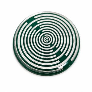 May include: A circular badge featuring a dark green background with a series of concentric silver rings that form a maze-like pattern. The design consists of multiple broken circular lines radiating from the centre towards the outer edge of the badge.