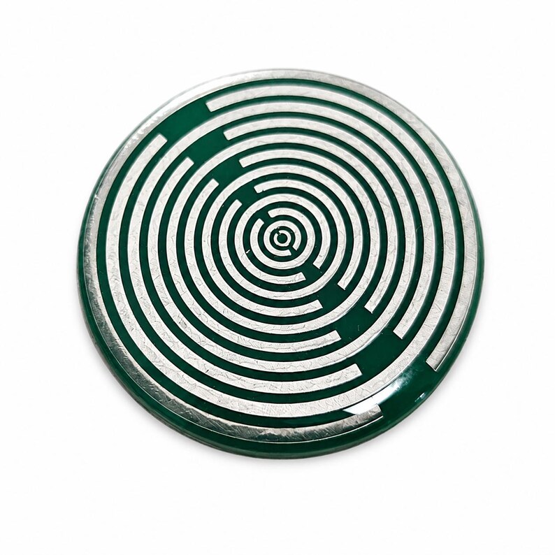 May include: A circular badge featuring a dark green background with a series of concentric silver rings that form a maze-like pattern. The design consists of multiple broken circular lines radiating from the centre towards the outer edge of the badge.