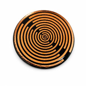 May include: A circular object featuring a metallic copper-coloured finish with a black concentric maze pattern. The design consists of multiple rings and breaks that form a labyrinthine structure across the flat, reflective surface of the disc.