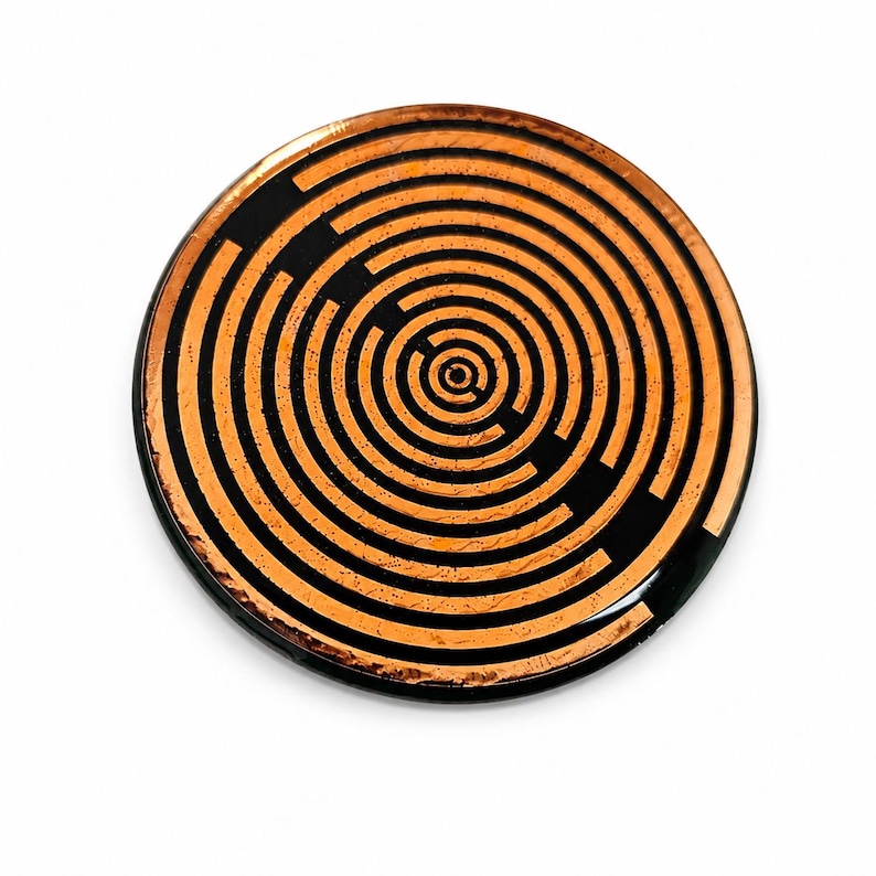 May include: A circular object featuring a metallic copper-coloured finish with a black concentric maze pattern. The design consists of multiple rings and breaks that form a labyrinthine structure across the flat, reflective surface of the disc.