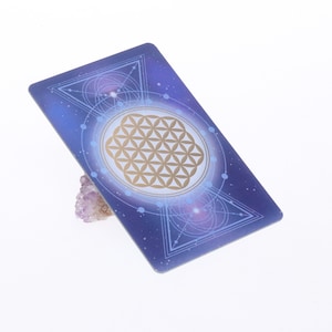 Puede incluir: Una carta de tarot con un diseño celestial, con un símbolo dorado de la Flor de la Vida sobre un fondo azul y morado. La carta está apoyada por un pequeño cristal de amatista de color púrpura claro. El diseño de la carta incluye formas y líneas geométricas.