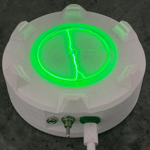 Puede incluir: Un dispositivo circular blanco con una superficie texturizada y seis pestañas elevadas en el perímetro. El centro presenta una luz circular verde brillante con un patrón irregular similar a un rayo. Un interruptor y un cable están conectados.