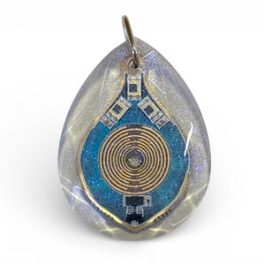 Può includere: Ciondolo a forma di goccia con anello argentato. Il ciondolo ha un esterno trasparente e glitterato che racchiude un design blu e oro con un centro a spirale. Il design ricorda un circuito stampato.
