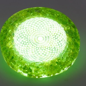 May include: A glowing, round, green decorative light fixture. The center features a geometric pattern, surrounded by a textured, green outer ring. The light emits a bright, white glow.