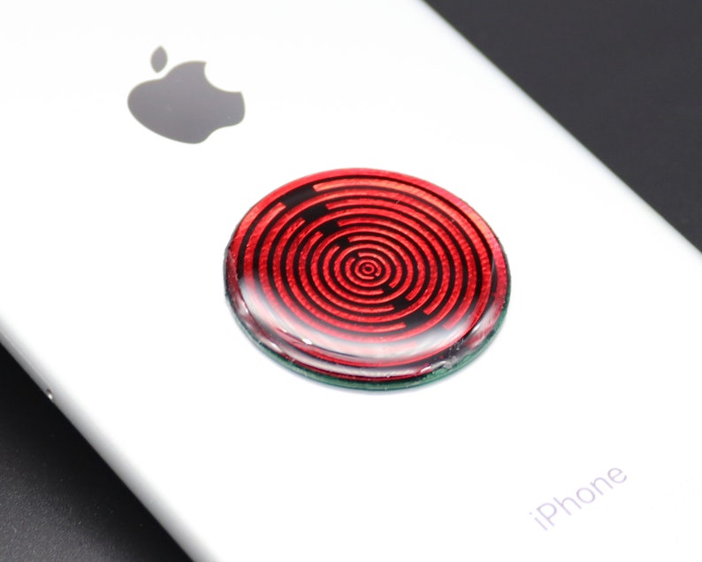 May include: A circular phone grip accessory featuring a glossy red surface with a concentric black spiral pattern. The disc is adhered to the back of a white smartphone, which displays the Apple logo and the word iPhone printed on the lower section.