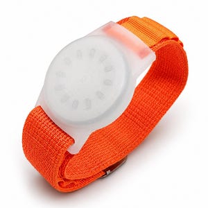May include: A bright orange elastic fabric wristband featuring a circular, translucent white plastic housing. The housing has a textured, circular face with a series of small, evenly spaced rectangular indentations arranged in a ring pattern.