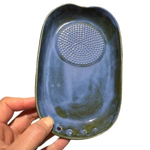 May include: A handmade, ceramic flower frog in a rectangular shape with a blue and green glaze. The frog has a textured, circular center for arranging flowers and three drainage holes. The item is held by a hand.