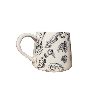 May include: A white ceramic mug featuring a repeating black ink illustration pattern of various sea creatures including a lobster, a squid, a fish, an octopus tentacle, and several types of seashells, complete with a curved side handle.