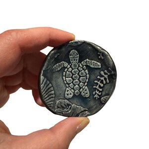May include: A round, ceramic coaster with a sea turtle design. The coaster is primarily dark blue with a lighter blue turtle in the center. The design includes sea plants and shells around the turtle. The coaster is held in a hand.
