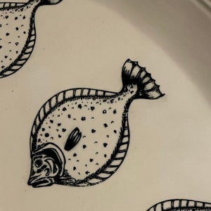 May include: A white ceramic plate featuring a black and white illustration of a fish. The fish has a spotted pattern and a detailed fin.