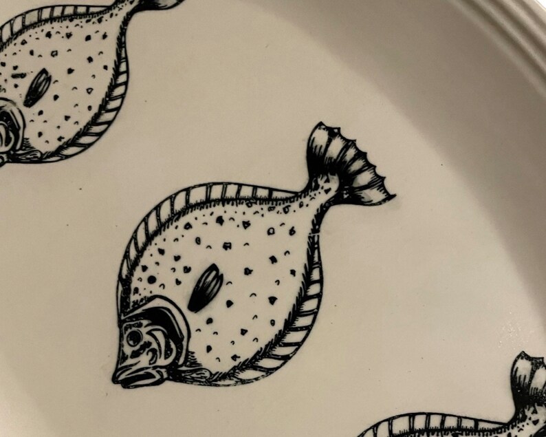 May include: A white ceramic plate featuring a black and white illustration of a fish. The fish has a spotted pattern and a detailed fin.