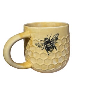 May include: A ceramic mug with a yellow honeycomb pattern and a black and white bee illustration. The mug has a curved handle and is approximately 4 inches tall.