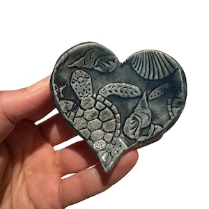 May include: A heart-shaped ceramic piece with a sea life design. The gray-blue piece features a turtle, seashells, and seaweed. The design is raised, creating a textured surface. The piece is held in a hand against a white background.
