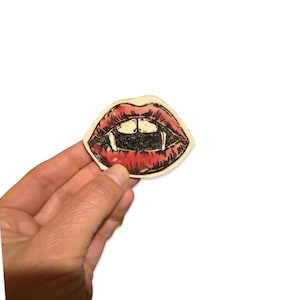 May include: A red and white sticker with a drawing of a mouth with fangs. The mouth is open and the fangs are visible. The sticker is on a white background.