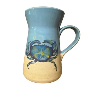 May include: A ceramic mug with a light blue top and a beige bottom. A blue crab illustration is painted on the front. The mug has a curved handle and a flared top.