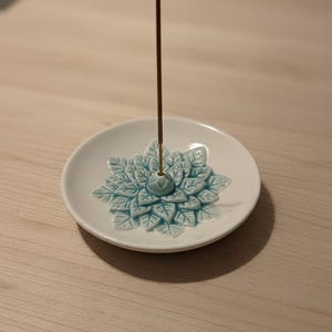 May include: White ceramic incense holder with a turquoise floral design. A brown incense stick stands upright in the center. The holder is round and sits on a light-colored wooden surface.