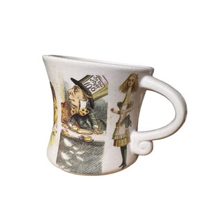 May include: White ceramic mug featuring illustrations from Alice in Wonderland. The mug depicts the Mad Hatter, Alice, and other characters. The handle has a unique, curved design. The mug is approximately 4 inches tall.