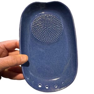 May include: A blue ceramic garlic grater with a textured grating surface in the center. The oval-shaped grater has a slightly curved design with three small holes at the base. The grater is held by a hand.