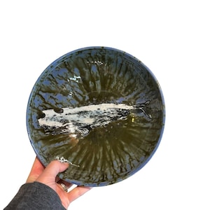 May include: A handmade ceramic bowl with a blue rim and a green and white interior. The bowl has a glossy finish and a unique, organic design. The bowl is round and appears to be approximately 10 inches in diameter.