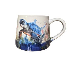 May include: A white ceramic mug featuring a watercolor illustration of a sea turtle swimming. The turtle has blue and orange markings, surrounded by blue and green splashes of color at the base. The mug has a curved handle on the right side.