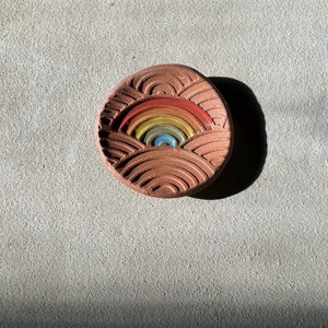 May include: A small, round, terracotta-colored ceramic dish with a rainbow design. The dish has a textured surface and is perfect for holding small items.