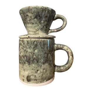 May include: A ceramic coffee pour-over set with a speckled green glaze. The set includes a mug with a handle and a matching pour-over cone. The mug has a white base.