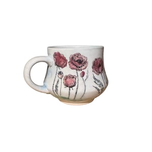 May include: A handmade ceramic mug with a white base and a curved handle. The mug is decorated with a watercolor-style floral design featuring pink poppies and green stems. The design wraps around the mug, creating a charming and artistic look.