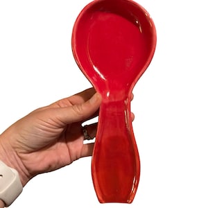 May include: A glossy, bright red ceramic spoon rest with a wide, circular bowl and a tapered handle section. The surface of the kitchen utensil features a smooth, reflective finish with subtle variations in the red glaze throughout the piece.