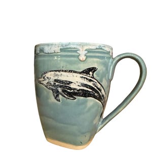 May include: A ceramic mug with a light blue glaze and a black and white dolphin illustration. The mug has a curved handle and a slightly textured rim. The mug is designed for hot beverages.