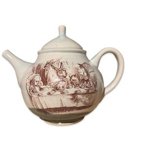May include: White ceramic teapot with a brown illustration of Alice in Wonderland characters having tea. The teapot has a lid and a handle.