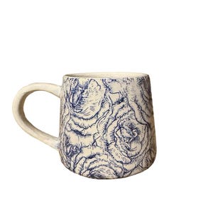 May include: Cream ceramic mug with a large handle. The mug features a blue floral pattern covering its surface, giving a textured look. Suitable for hot or cold drinks. The mug's design is reminiscent of a botanical illustration.