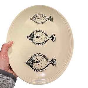 May include: A white oval platter with three black and white fish illustrations. The fish are facing to the right and have a spotted pattern.