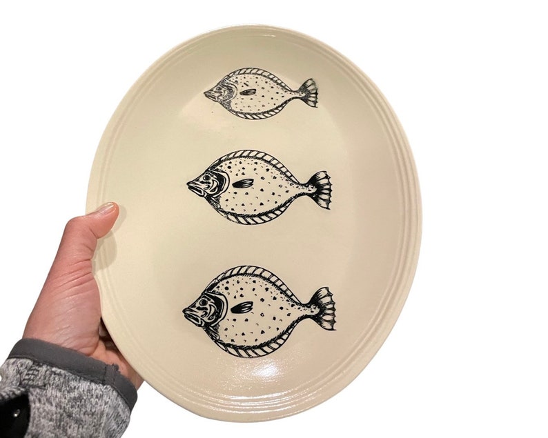 May include: A white oval platter with three black and white fish illustrations. The fish are facing to the right and have a spotted pattern.