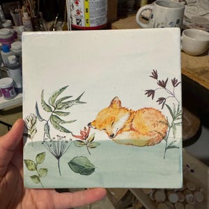 May include: Square ceramic tile featuring a watercolor illustration of a sleeping fox. The fox is orange and white, resting among green and brown foliage. The tile has a white background with a light green border at the bottom.