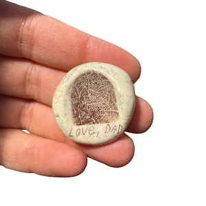 May include: A small, round, off-white ceramic keepsake with a detailed fingerprint impression in brown. The words "Love, Dad" are handwritten in black ink below the fingerprint. The keepsake is held in a hand against a white background.