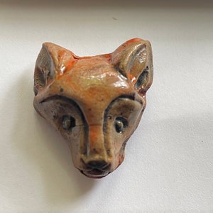 May include: A ceramic fox head sculpture with brown and orange glaze. The fox has a detailed face with small eyes and a pointed nose.