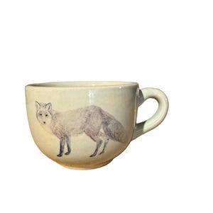 May include: Cream-colored ceramic mug with a gray fox illustration. The fox is depicted in a side profile, with detailed fur and a bushy tail. The mug has a rounded shape and a curved handle.