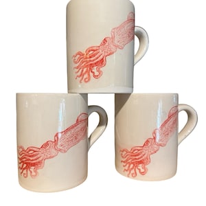 May include: Three white ceramic mugs stacked, each featuring a red illustration of a squid. The squid design wraps around the mug's body. The mugs have a simple, cylindrical shape with a curved handle.