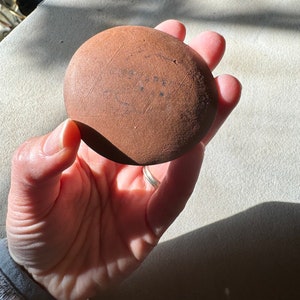 May include: A round, brown, terracotta-colored clay pot with a smooth surface. The pot has a slightly uneven shape and is marked with a series of lines and dots.