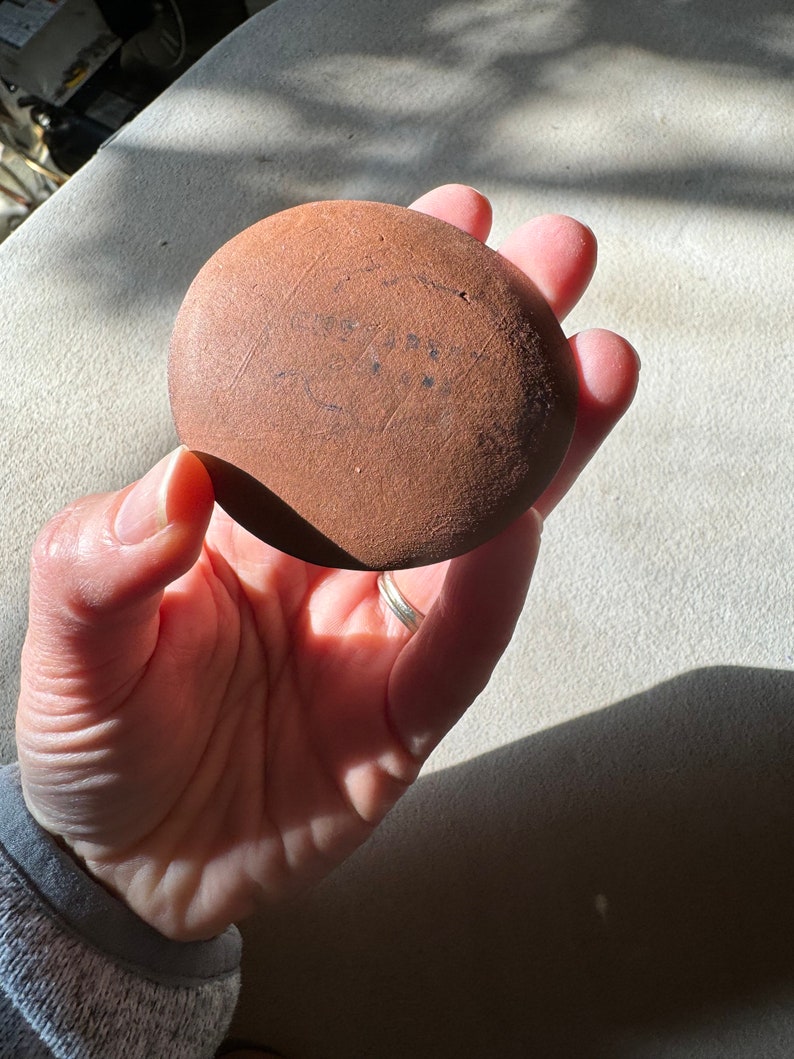May include: A round, brown, terracotta-colored clay pot with a smooth surface. The pot has a slightly uneven shape and is marked with a series of lines and dots.