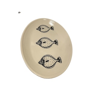 May include: A white oval ceramic serving platter with a black outline of three fish. The fish are facing to the right and have a spotted pattern.
