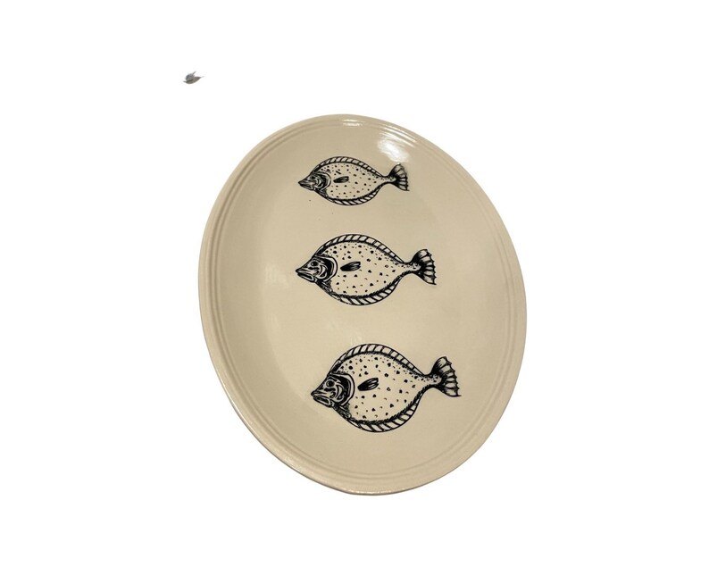 May include: A white oval ceramic serving platter with a black outline of three fish. The fish are facing to the right and have a spotted pattern.