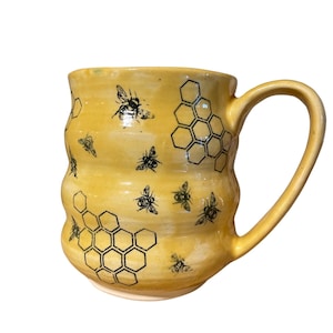 May include: A yellow ceramic mug with a wavy design, featuring black illustrations of bees and honeycomb patterns. The mug has a large handle and a capacity suitable for hot beverages. The overall design evokes a natural, honey-themed aesthetic.