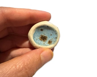 May include: A small, round ceramic dish with a light blue glaze and brown speckles. The dish is slightly raised in the center.