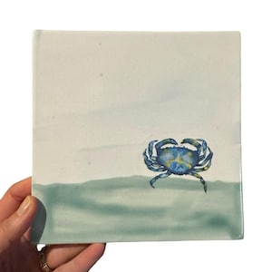 May include: Square ceramic tile featuring a watercolor painting of a blue crab on a light blue and green background. The crab has yellow accents. The tile is held by a hand.