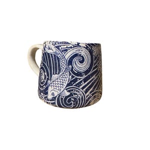 May include: A ceramic mug with a white handle and a blue and white koi fish design. The mug features a repeating pattern of koi fish and swirling water. The mug is a standard size for enjoying hot beverages. The design evokes a sense of calm.