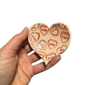 May include: A heart-shaped ceramic dish with a white background and red hearts printed with various messages, including "Hugs", "Sweet", "Love", "Forever", "Be Mine", and "Kisses".