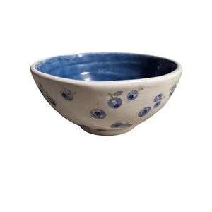 May include: A ceramic bowl with a white exterior and a blue interior. The bowl is decorated with hand-painted blue blueberry designs. The bowl is a small size, perfect for serving snacks or small portions.