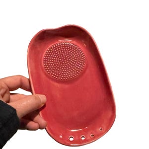 May include: An oval-shaped, rose-colored ceramic grater plate with a textured, circular grinding surface in the center. The plate features a smooth, glossy finish and includes four small, circular drainage holes along the bottom edge.