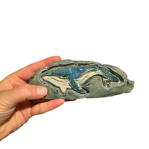 May include: A ceramic whale sculpture in shades of blue and gray. The whale is depicted in a detailed, artistic style, with a textured surface. The sculpture is held in a hand against a white background.
